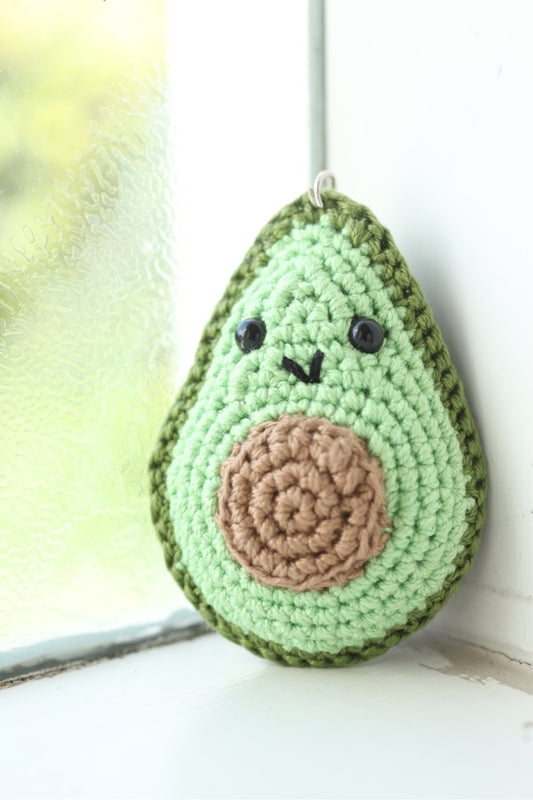 A crochet keychain in the shape of an avocado with a smiling face, predominantly green in color with a brown centre 'stone'