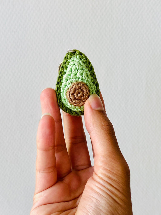 A crochet keychain in the shape of an avocado, with green outer layers and a brown 'stone' in the centre