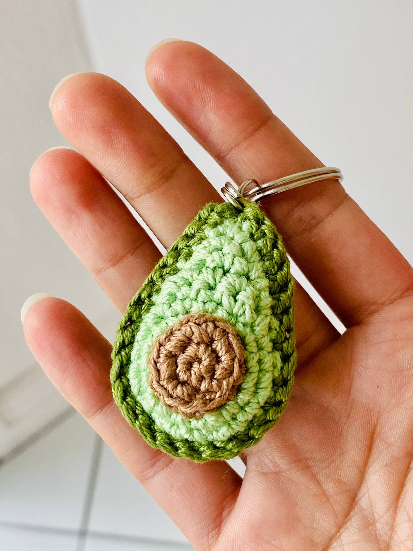 A crochet keychain in the shape of an avocado with green outer layers and a brown 'stone' laying flat on a person's hand