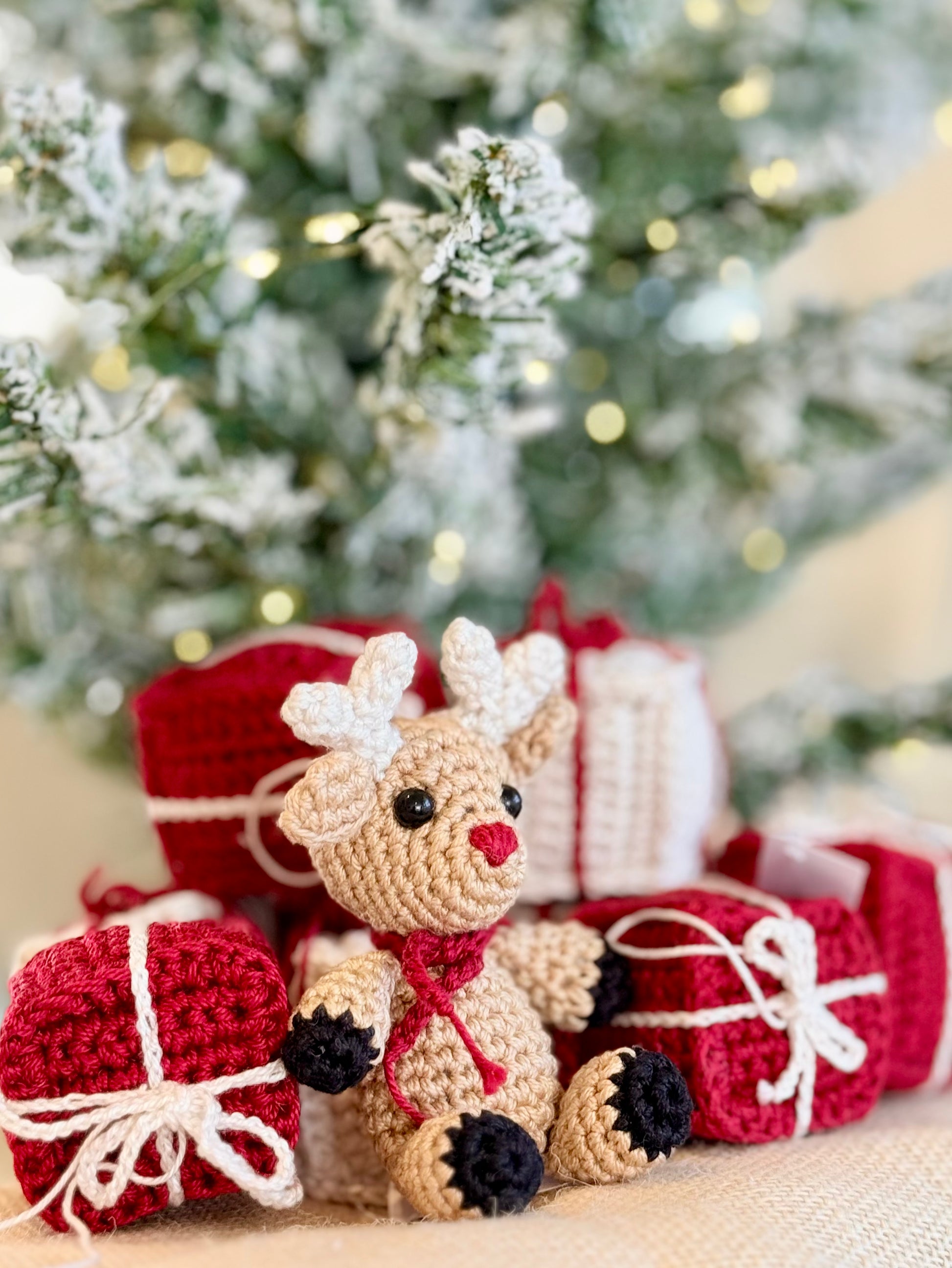 A crocheted reindeer sits in front of wrapped Christmas presents, with a blurred festive tree in the background. The reindeer is beige with white antlers, a red nose and scarf, and black paws.