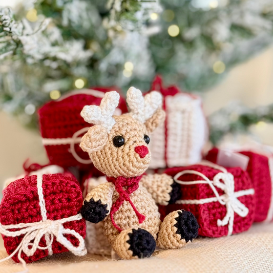 A crocheted reindeer sits in front of wrapped Christmas presents, with a blurred festive tree in the background. The reindeer is beige with white antlers, a red nose and scarf, and black paw pads, surrounded by red and white gift boxes with white string bows.