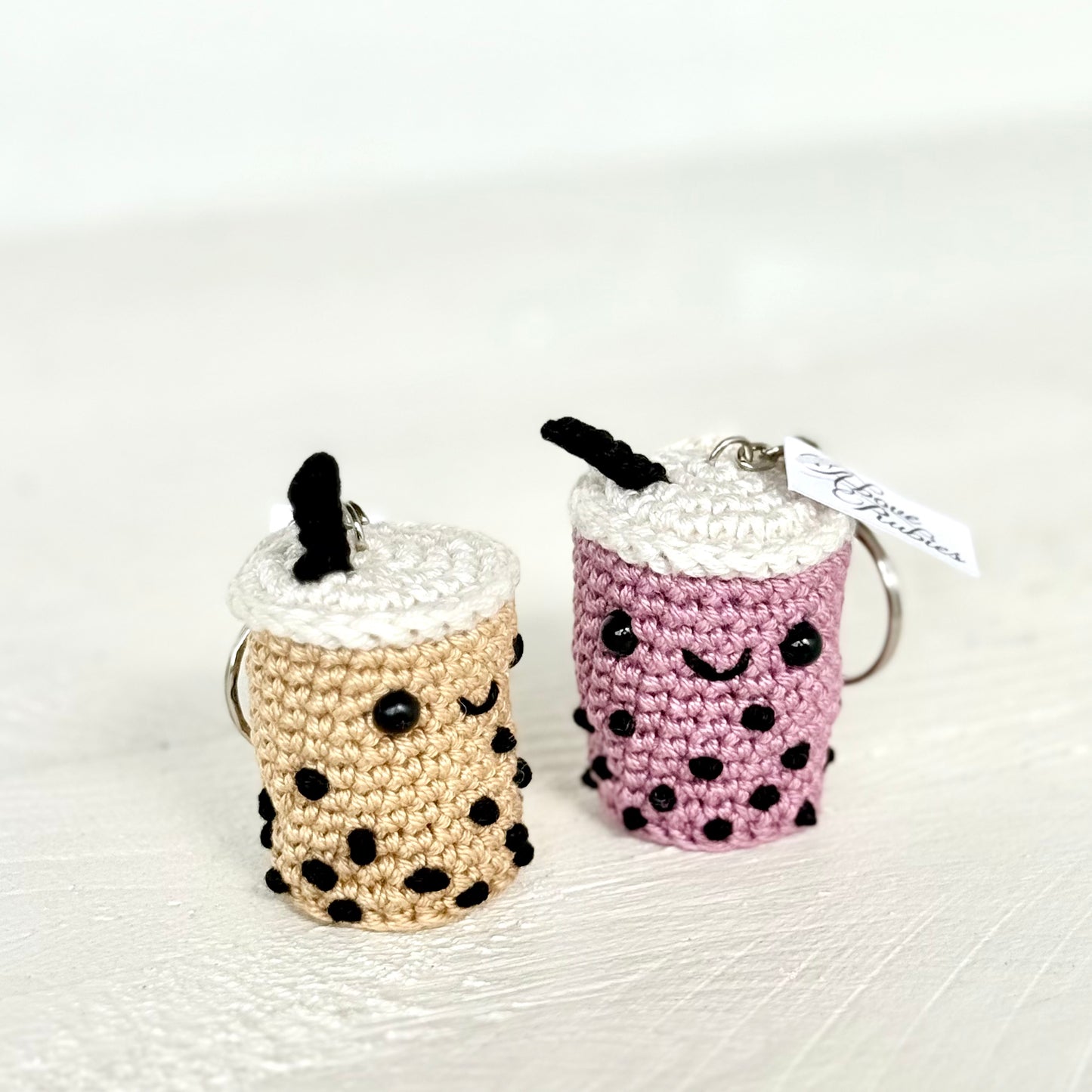 Two crocheted boba tea keychains on a white textured surface. One keychain is beige with black boba dots and a black straw, and the other is purple with black boba dots and a black straw.
