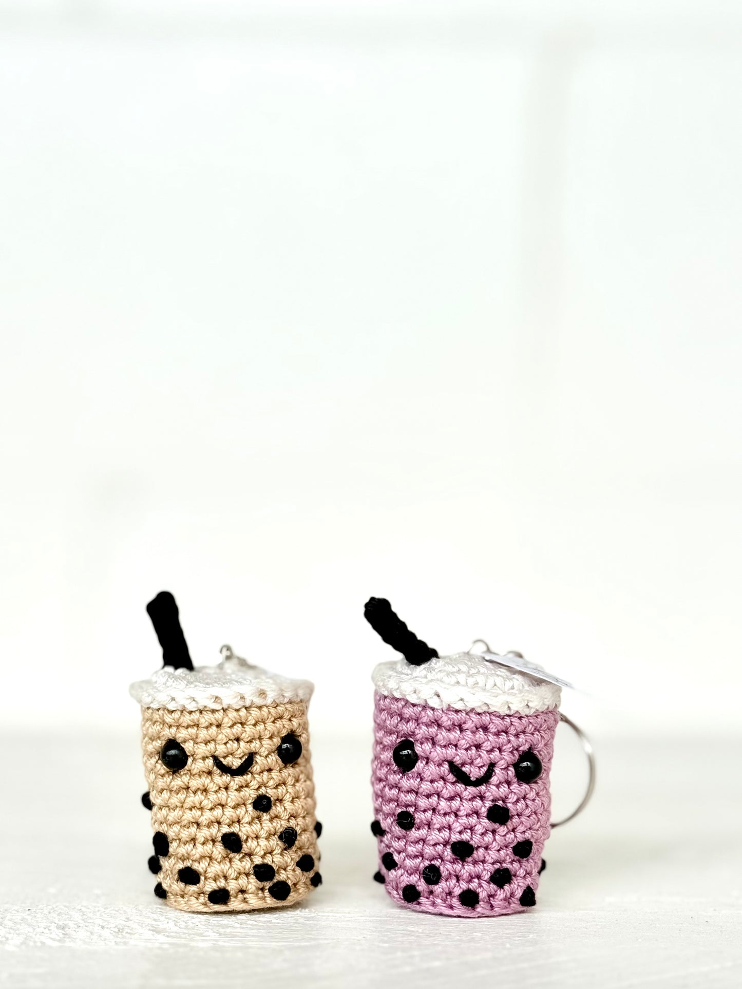 Two small crocheted boba tea keychains on a white surface against a white background. One keychain is beige with black polka dots and a black straw, and the other is purple with black polka dots and a black straw.