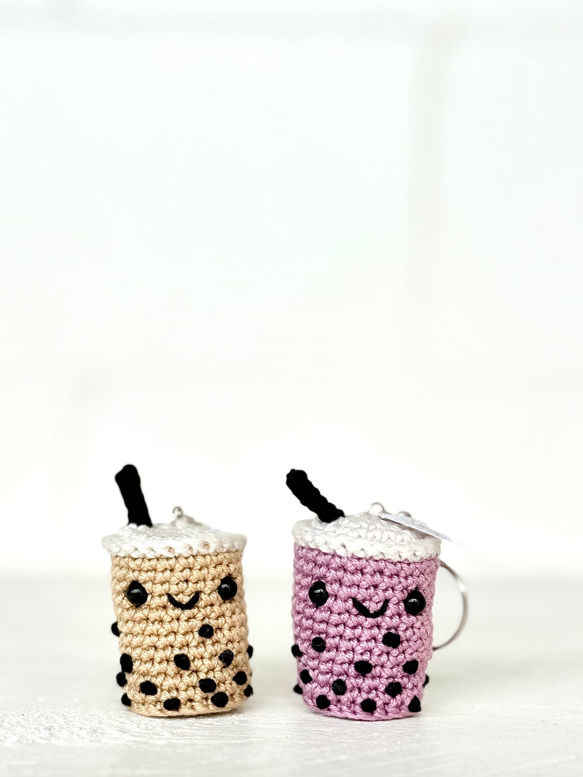 Two small crocheted boba tea keychains on a white surface against a white background. One keychain is beige with black polka dots and a black straw, and the other is purple with black polka dots and a black straw.