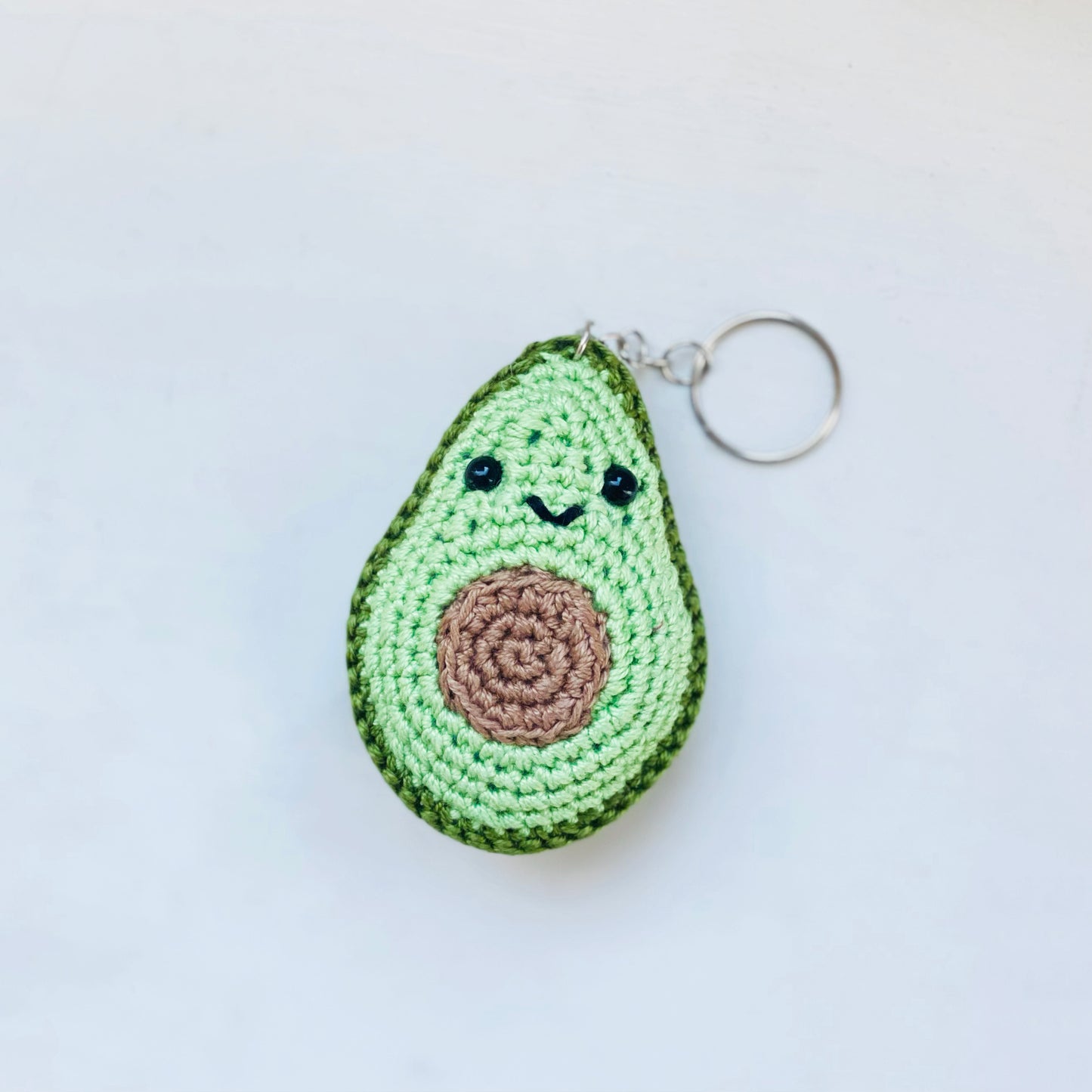 A crochet keychain in the shape of an avocado with a smiling face, predominantly green in color with a brown centre 'stone'