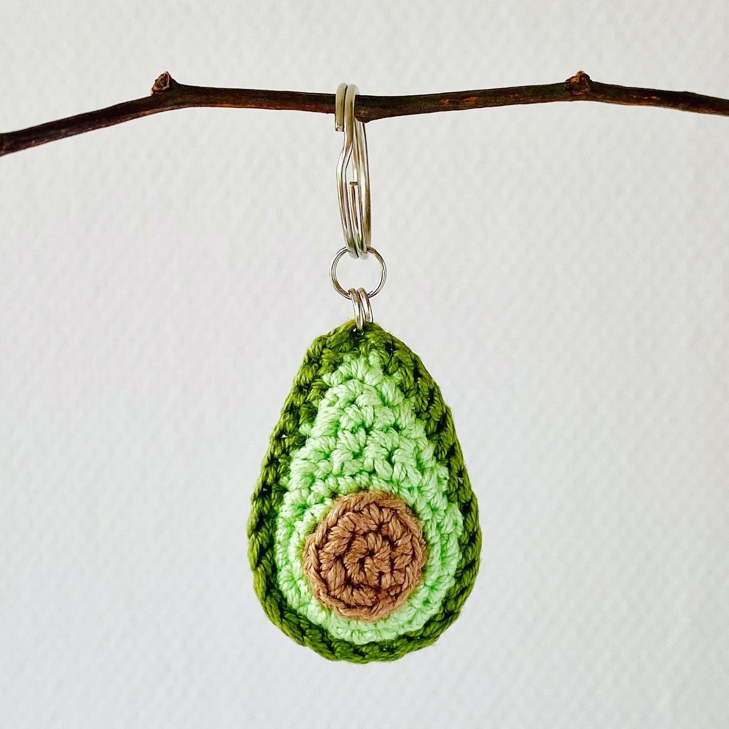 A crochet keychain in the shape of an avocado, with green outer layers and a brown 'stone' in the centre hanging from a metal ring.